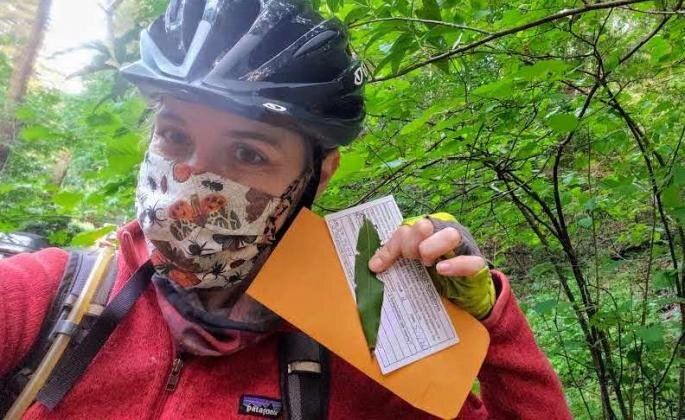 Someone wearing a mask, bicycle helmet, and outdoor gear while holding/posing with some paperwork and a leaf