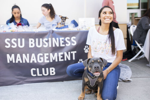 A student smiling and posing with a dog in front of a table display featuring a banner that reads "SSU Business Management Club"