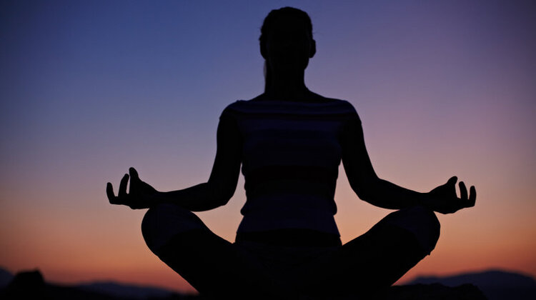 The silhouette of someone in a meditative pose in front of an orange, purple, and blue sunset