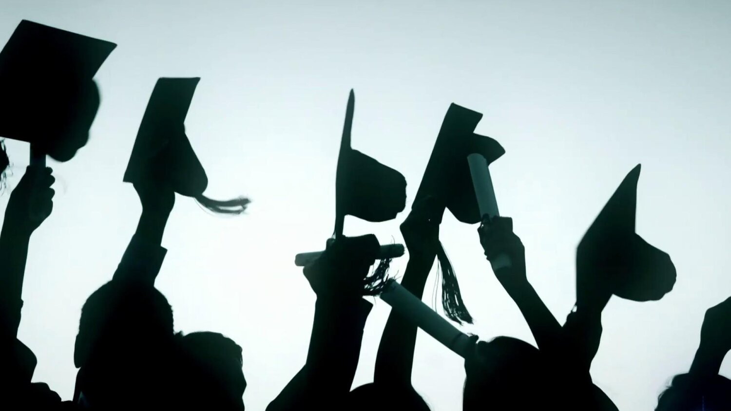 The silhouette of graduates tossing their graduation caps in the air in front of a pale blue background