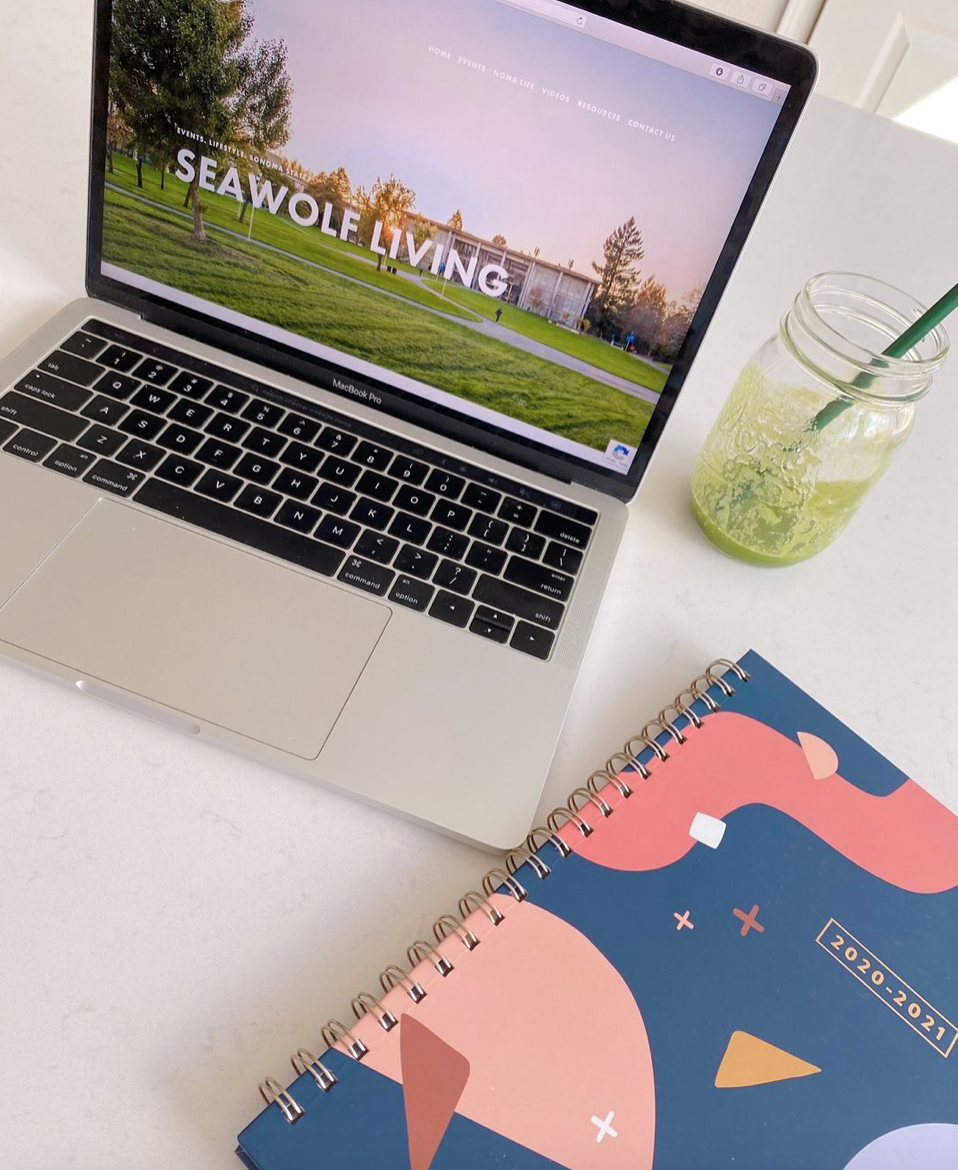 A colorful planner, a green iced drink in a glass jar with a green straw, and an open laptop with the words "Seawolf Living" on the screen