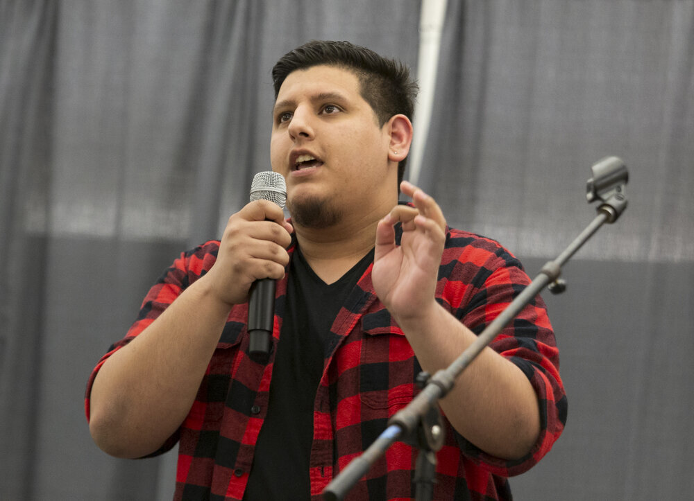 A speaker wearing a red and black plaid flannel and speaking into the microphone in one of their hands while making a gesture with their other hand