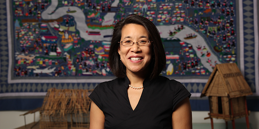 A portrait of Dr. Erika Lee smiling in a black shirt in front of artwork and artifacts 