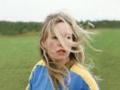 A person with long blond hair blowing in the wind and a blue and yellow shirt standing in a green field