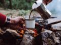 Somebody pouring someone a mug of coffee in front of a campfire