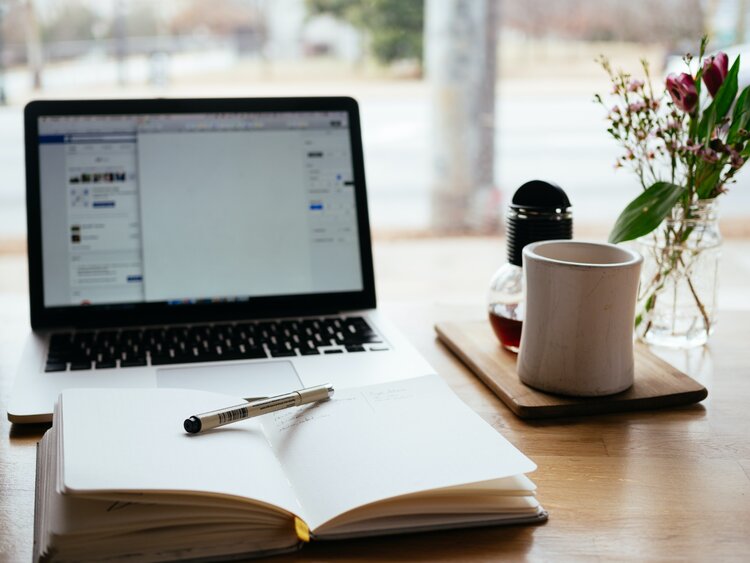 A well-lit desk with an open laptop, open notebook, pen, coffee cup, and small arrangement of flowers 