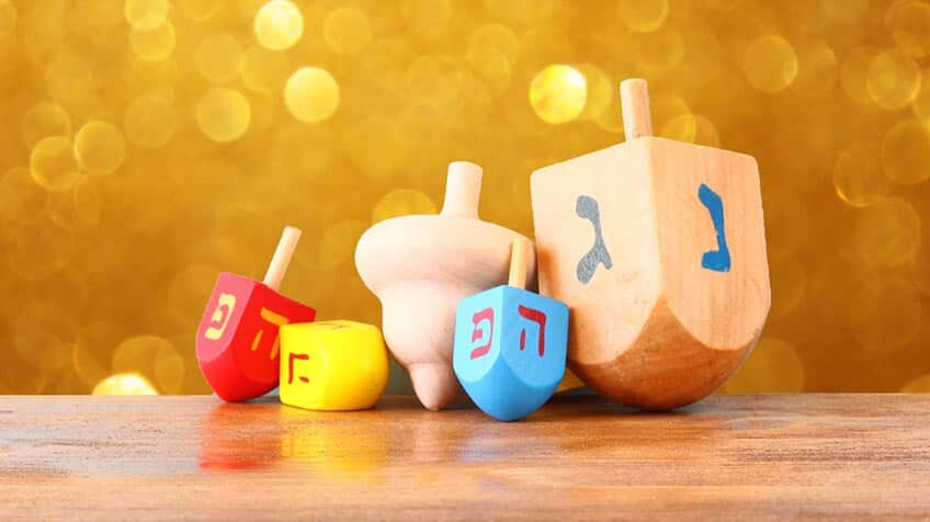 Five red, yellow, and blue dreidels on a wooden surface in front of a gold bokeh background