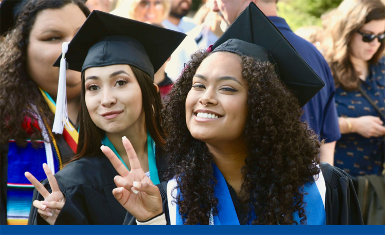 Two graduates wearing graduation caps and gowns smile and pose with finger peace signs 