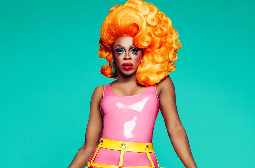 Portrait of Honey Davenport with orange hair, colorful makeup, and a pink latex dress on in front of a teal background