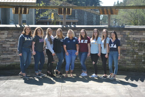 A line of nine sorority members smiling while wearing different sorority sweatshirts in an outdoor setting