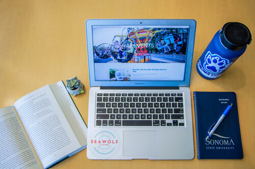 A tabletop featuring a thermos featuring a Seawolf paw, a Sonoma State University notebook, a pen, an open book, and an open laptop with a Seawolf Living sticker on it