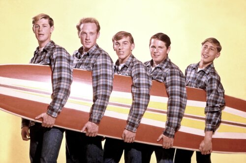 The five band members of The Beach Boys in a line wearing matching flannels while holding a singular surfboard