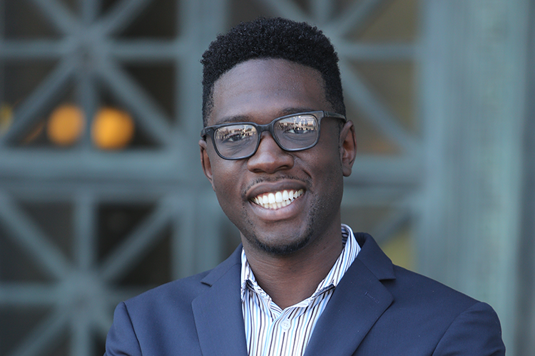 A portrait of Travis Bristol smiling while wearing a navy blazer with a striped button-up shirt underneath