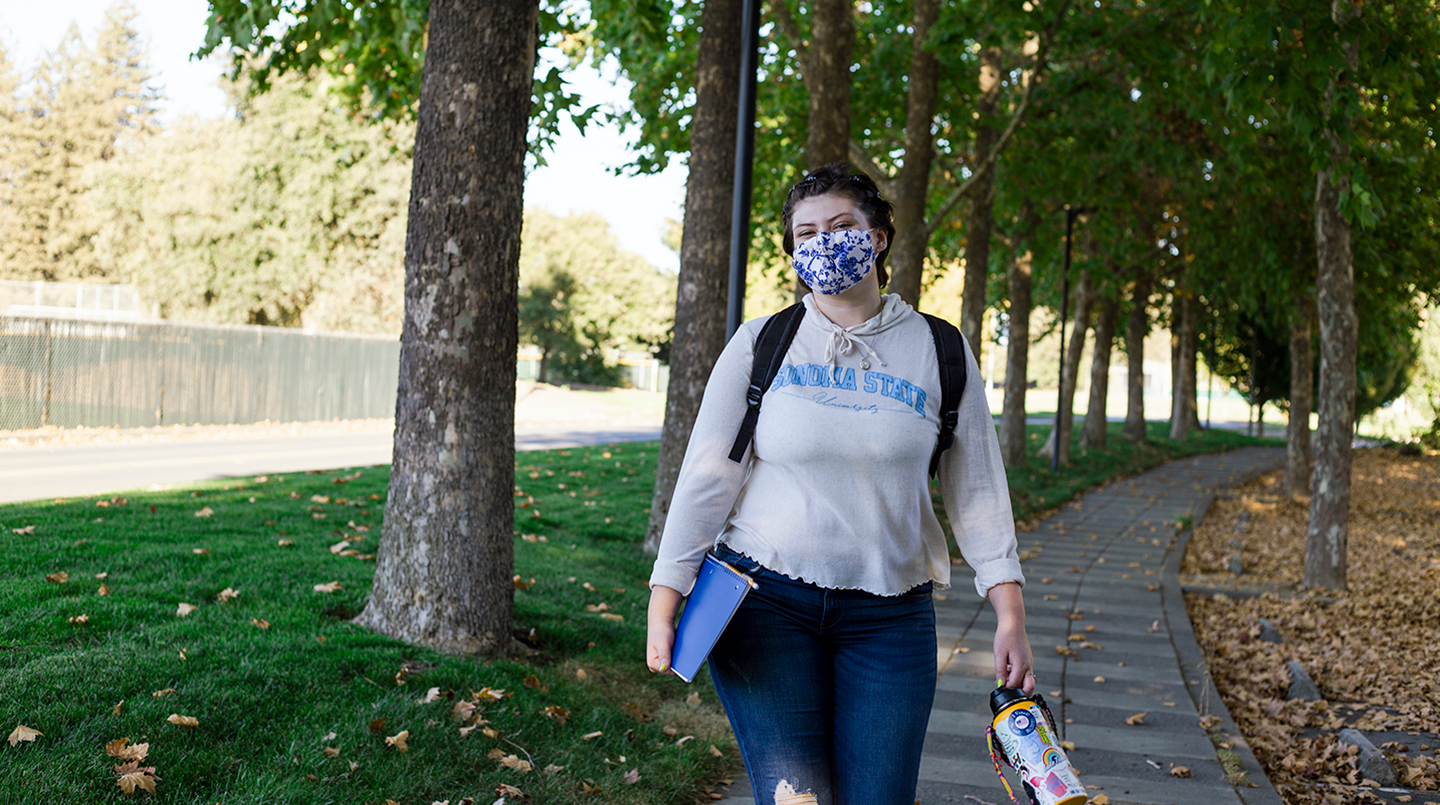 Student holding books and water bottle walks down tree-lined sidewalk 