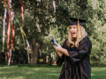 A graduate with a surprised expression popping a confetti party popper while wearing a graduation cap and gown 