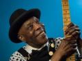 A portrait of Buddy Guy smiling and posing with his instrument 