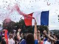 a person holding a red smoke bomb in a crowd of people with another person holding the French flag and confetti falling from the sky