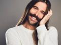Portrait of Jonathan Van Ness smiling while wearing white sweater in front of a faded grey background