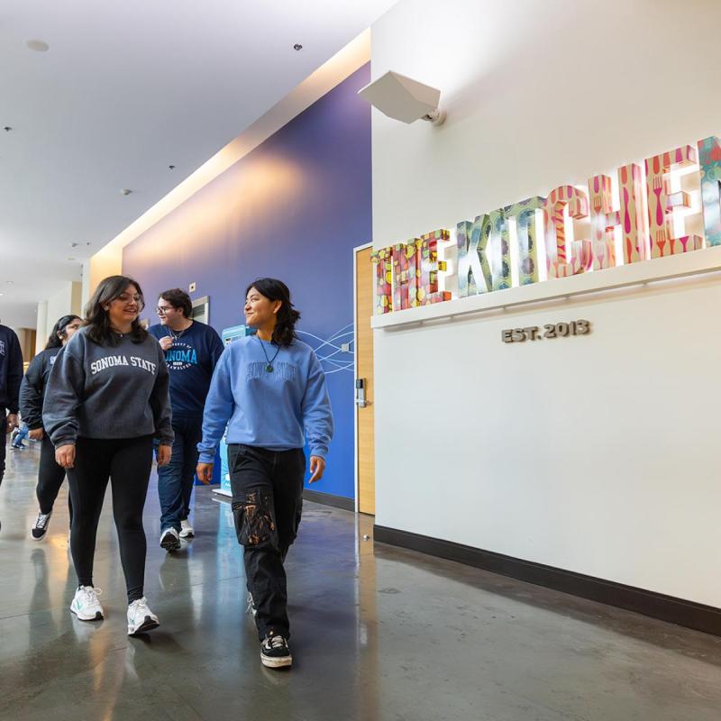 Four students walking in a hallway with the sign The Kitchens behind them