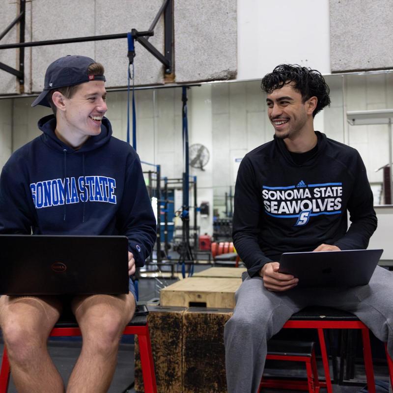 Two people sitting and wearing Sonoma State shirts and on computers looking at each other